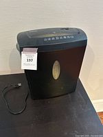 Front and side view of black Staples paper shredder sitting on black surface with power cord visible, showing transparent oval window for shredded paper.