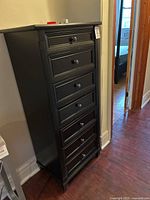 Tall black wooden dresser with 7 drawers against a wall on hardwood floor.