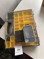 Plastic toolbox with yellow compartments and clear lids showing screws and screwdriver bits inside on a white surface by a wall.