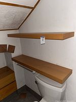 Photo showing two floating wooden shelves mounted on a white wall above toilet and next to stairs. Shelves are rectangular with a smooth medium brown wood finish.
