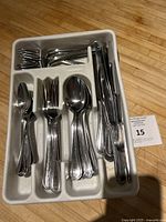 Photo of multiple silver utensils including forks, knives, teaspoons, tablespoons organized in a white plastic tray on a wooden floor.