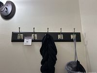 Black wall-mounted hook rack with five silver metal hooks attached, with a light fixture visible on upper left and some fabric hanging on hooks.