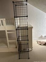 Full view of the black wire shelving unit upright beside white furniture, showing the five connected cubical compartments.