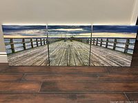 Photo of three square canvas panels arranged horizontally to form a continuous image of a wooden pier leading out to water and distant horizon under a cloudy sky