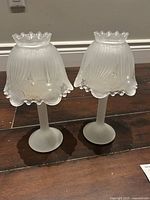 Pair of vintage frosted glass candle lamps with scalloped bell-shaped frosted glass shades and ruffled clear glass trim on a wooden surface