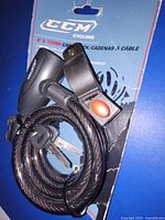 CCM branded black cable bicycle lock coiled on original blue packaging, showing lock body with red button and two keys inserted on top side.