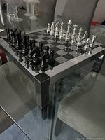 Glass chess board with black and silver metal chess pieces arranged at start position on a glass table.