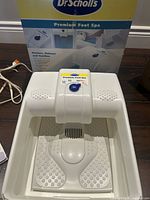 Electric foot spa unit showing white plastic basin with textured footpads, central massager, and control knob on the top labeled Premium Foot Spa by Dr Scholl's.