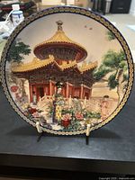 Front of the decorative plate showing a detailed, colorful scene of the Pavilion of 1000 Springs with traditional figures and ornate architecture under a gold-tone rim.