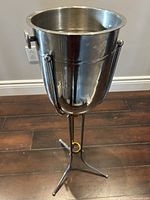 Photo showing a tall stainless steel ice bucket with a handle attached on the sides. The bucket sits on a three-legged stand and reflects surroundings.