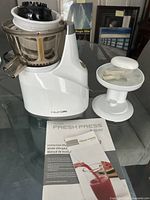 Photo of white Hurom Fresh Press juicer disassembled with components and user manual on glass table.
