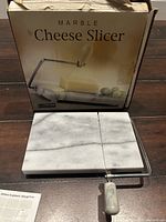 Photo showing marble cheese slicer with its original packaging box. The white marble base contrasts with a metal wire cutting arm and marble handle.