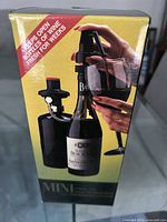 Boxed mini wine preserver and pour spout device with black air pump, shown attached to a wine bottle on packaging.