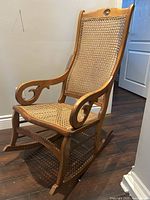 Photo of a wooden rocking chair with woven wicker seat and backrest, curved armrests, and rocker base, placed on wooden floor near a wall and door.