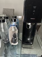 Black SodaStream sparkling water maker with textured front and button controls, next to 4 clear plastic carbonating bottles with black caps, one still in packaging.
