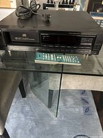 Front and top view of a black Sony 10 disc automatic CD changer placed on a glass surface with accompanying remote control in front.