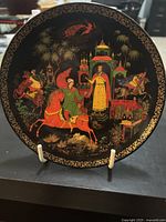 Front view of the black porcelain plate with detailed colorful painted scene and gold ornate border. The painting shows a royal figure, horse riders, and architectural elements.