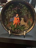 Front view of a decorative plate with intricate gold border and a colorful illustration of Tsarevich Ivan with a grey wolf and birds in a forested setting.