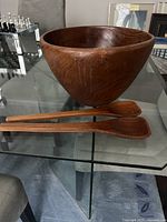 Photo of large wooden bowl with two wooden utensils placed on a glass table, showing the side profile and texture of the bowl and utensils.
