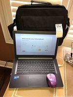 Photo showing Asus Chromebook laptop open with welcome screen displayed, purple wireless mouse on touchpad, and black carrying bag standing upright behind