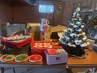 Photo showing ceramic tree, coasters, napkins, plates, cups, boxed cake stand, and string lights box on table in basement setting.