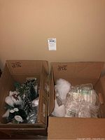 Two open cardboard boxes containing Christmas Village accessories including trees, fake snow, stands, and packaged lights.