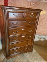 Front view of the brown MDF dresser with seven drawers and metal handles, showing detailed carved edges and metal pulls.