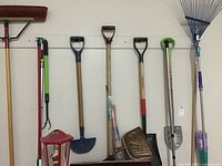 Photo showing garden tools hung on the wall: two digging shovels with wooden handles and metal heads, a red snow shovel with a wide plastic blade, large garden rake with metal tines, red broom with wooden handle.