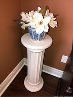 Full view of the white ceramic pedestal topped with a blue and white ceramic flower pot containing white faux flowers in a corner of the room.