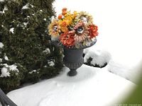 Black heavy metal planter on a pedestal base in snow with colorful faux flowers inside.