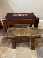Two antique wooden stools shown side by side on carpeted floor against white wall. Small foot stool with rough aged wood in front, larger wood framed stool with dark needlepoint floral seat behind.