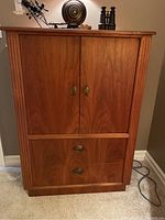 Front view of the wardrobe cabinet showing two doors on top with oval brass handles and two drawers underneath with matching brass pulls. The wood veneer finish is medium brown with visible grain.