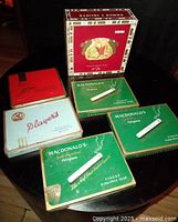 Five vintage cigarette tins and one Romeo Y Julieta cigar box on round wooden table, showing brand details and varying colors.