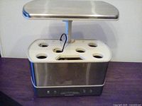 Front view of AeroGarden Harvest Elite hydroponic garden showing six planting holes and digital control panel on base with stainless steel cover and grow light overhead.