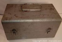 Exterior of the vintage metal carrying case with a handle and latch closures, showing wear and patina on the surface.