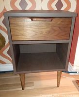 Front view of vintage nightstand showing walnut drawer front, painted brown body, and wooden legs.