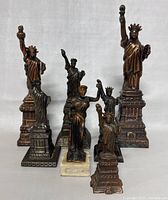 Group photo showing six metal decorative statues: five Statue of Liberty replicas alongside one vintage metal female figure statue on a white marble base, displaying their relative sizes and finishes.