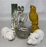 Front view of all five items: yellow Avon glass budgie perfume bottle, ceramic bud vase with dog and birds, glass swan figurine, and two white ceramic pigs.