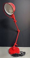 Red metal adjustable folding desk lamp standing upright showing shade, arm joints, weighted circular base, and power cord coiled at base.