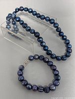 Photo showing the necklace coiled on a stand and the bracelet laid flat beside it, both with peacock-colored pearls and silver clasps.