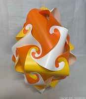 Photo showing the assembled spherical puzzle pendant lamp with orange, yellow, white, and peach curved puzzle pieces creating an intricate design, illuminated by ambient light.