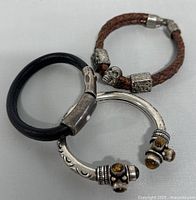Three bracelets displayed together including silver plated tiger eye cuff, brown braided leather skull bracelet, and black leather bracelet.