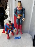 Photo of two Superman action figures placed side by side; smaller figure approximately 6 1/4 inches tall and larger figure approximately 11 1/2 inches tall, both with iconic blue and red costumes and capes.