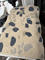 Beige rug with dark gray and taupe leaf and floral patterns shown spread out on a surface.