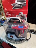 Portable Hoover CleanSlate spot cleaner front and side view with red and gray details, transparent water compartments, hose, power cord, and original red box in background.