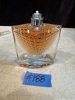 Front view of clear diamond-patterned glass perfume bottle containing amber liquid, metal spray nozzle visible but cap is missing.