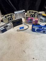 Lot of 11 cameras arranged on surface including vintage beige Easy Shot film cameras, digital compact cameras, disposable multicolor wrapped Fujifilm film camera, and blue Polaroid instant camera.