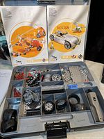 Top view of open plastic carrying case showing organized metal parts, wheels, screws, and motor attachment with two instruction manuals behind.