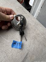 Image showing a silver discus padlock marked 'Abus' held in hand with three attached black keys and a small blue tag marked #03.