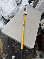 Full view of the Rubbermaid commercial floor cleaning tool with extended yellow adjustable handle on a grey table outdoors with snow around.